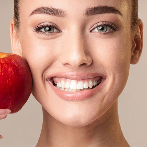 Eine Nahaufnahme zeigt das Gesicht einer jungen Frau, die einen roten Apfel an ihre Wange hält und breit lächelt. Ihre hellen Augen schauen direkt in die Kamera und ihre weißen Zähne sind gut sichtbar.